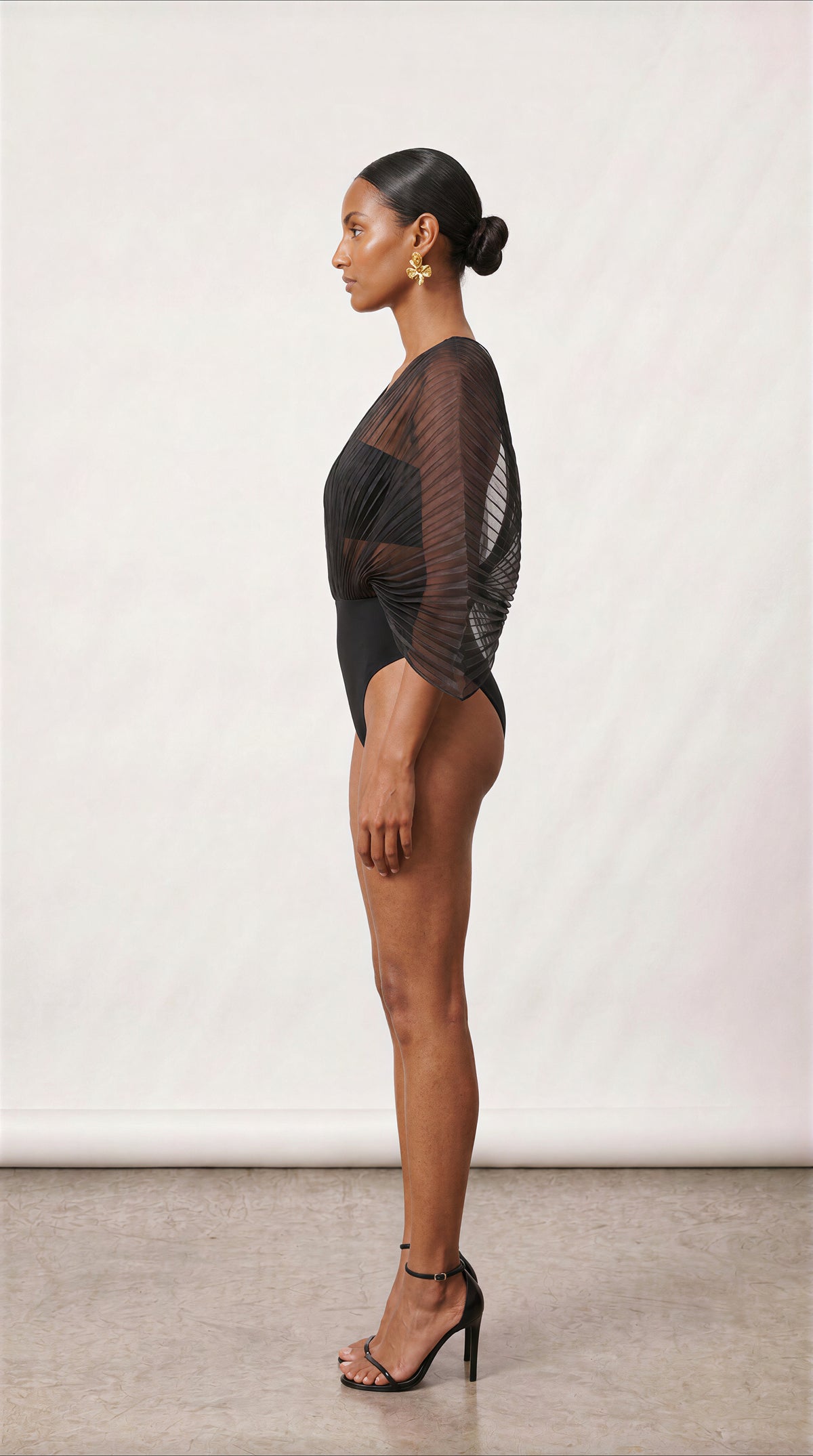 A woman stands in profile wearing a sheer black long-sleeve bodysuit with a fitted bodice and high-cut legs, paired with black strappy high heels, against a plain light studio backdrop.