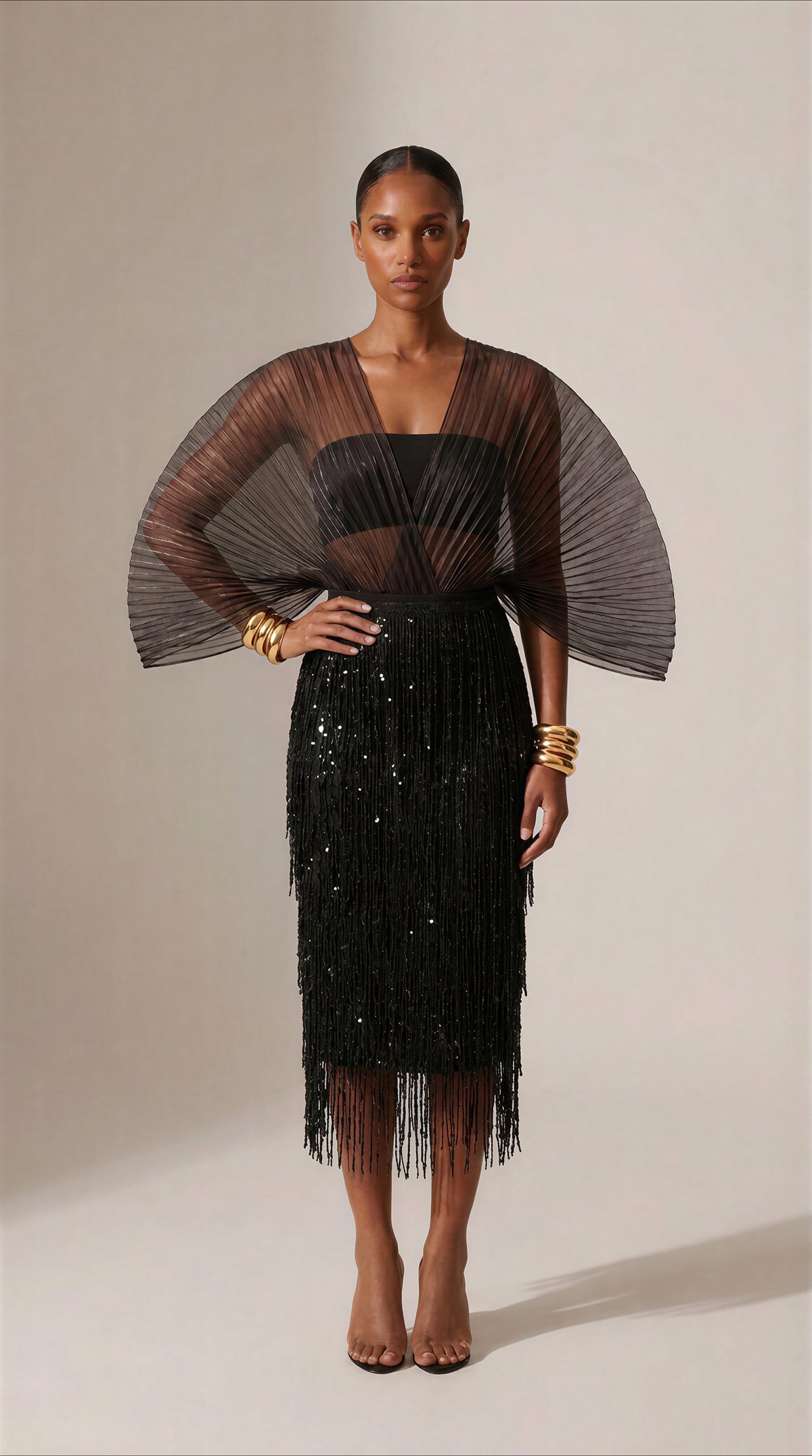 A model stands against a warm beige studio background wearing a black embellished fringe midi skirt with a sheer black mesh cape-style top overlay and gold bracelets.